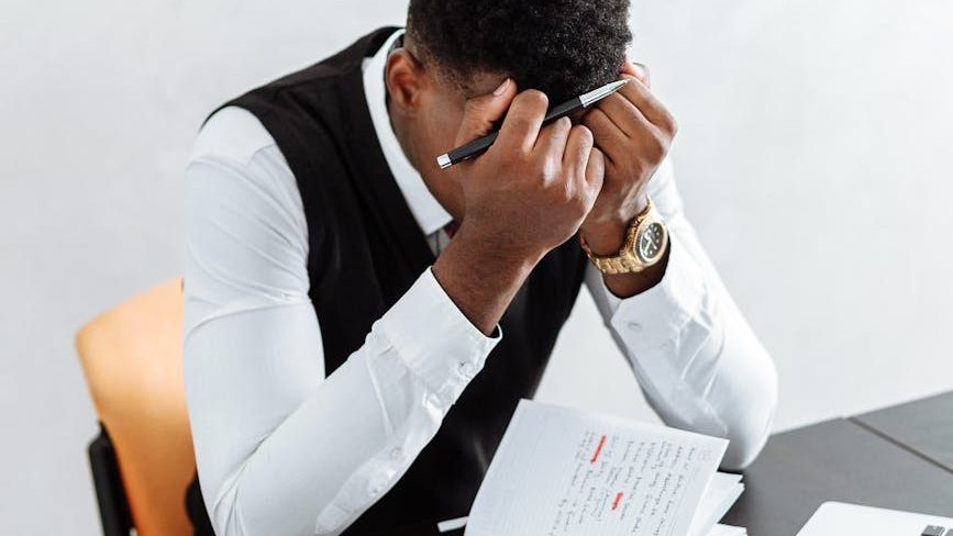 Person sitting at cluttered desk with papers and folders, looking overwhelmed and stressed.