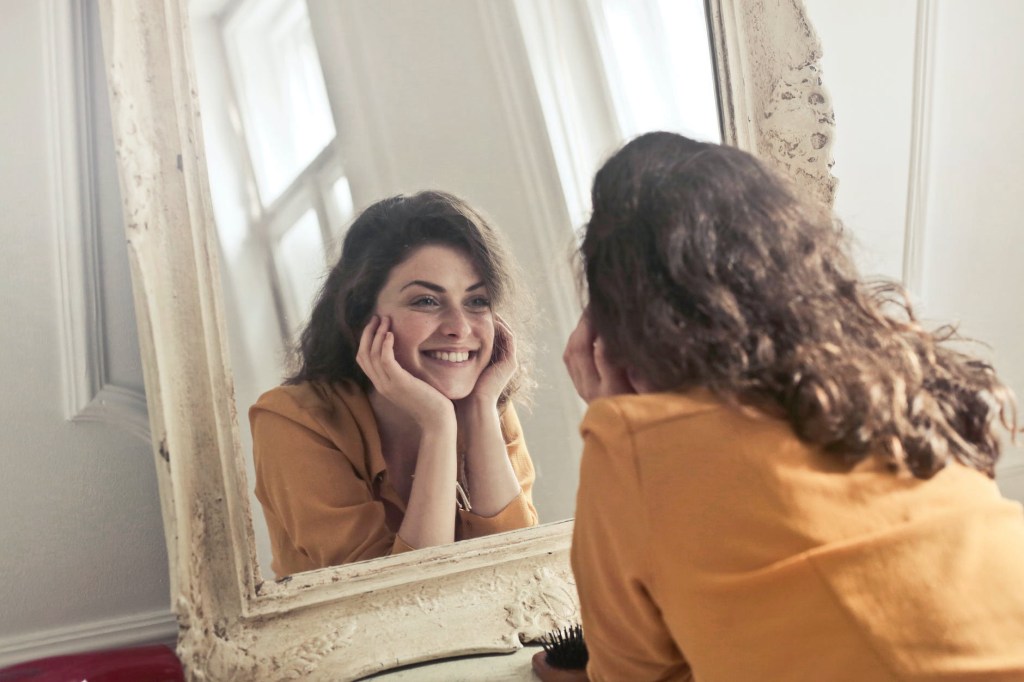 A person holds a mirror and smiles at their reflection, practicing self-love and acceptance. Jennifer Adair LPC, Onesta Therapy Co.